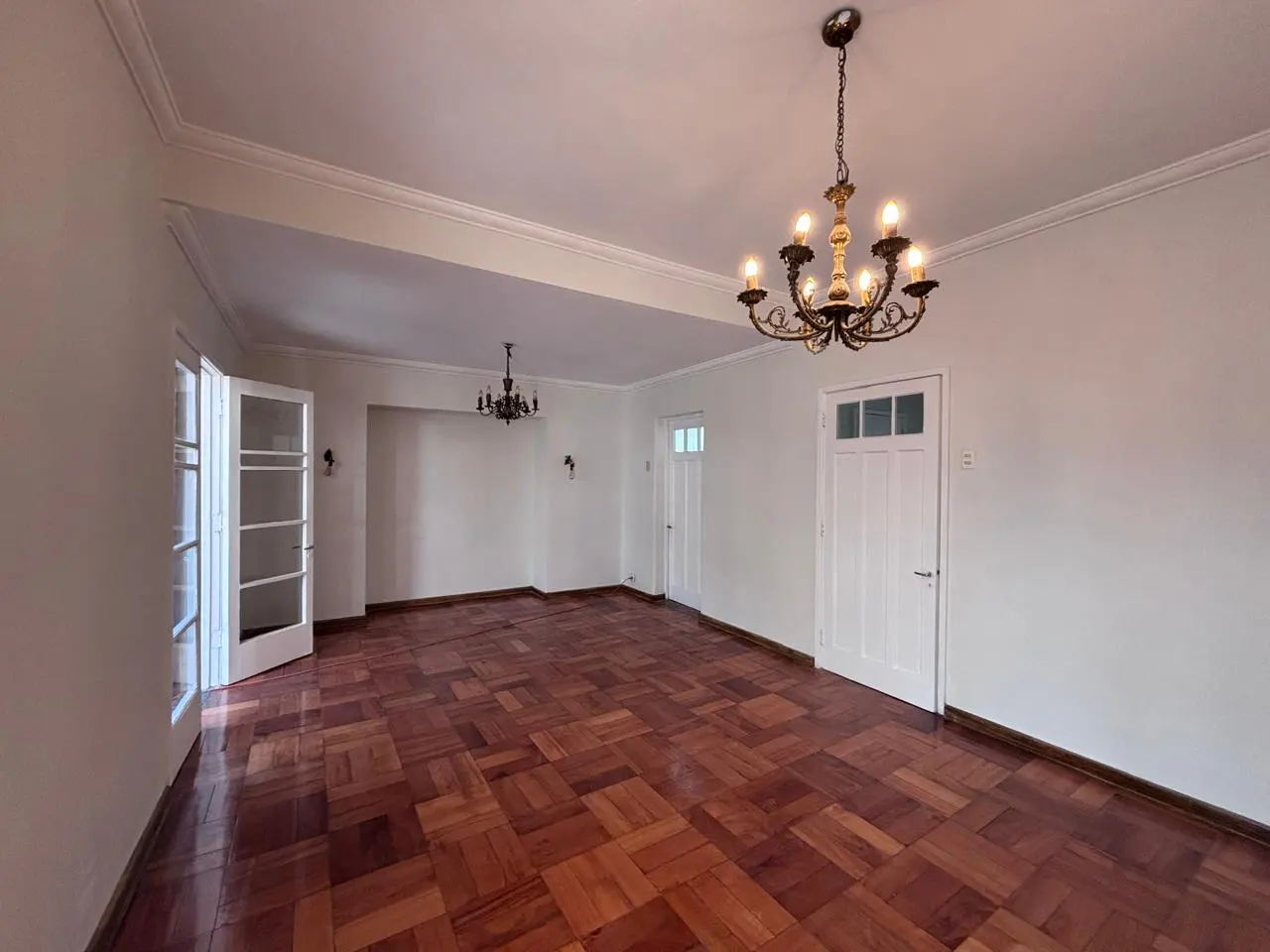 Empty room with parquet floors, white walls, and two chandeliers. A glass door is open on the left.