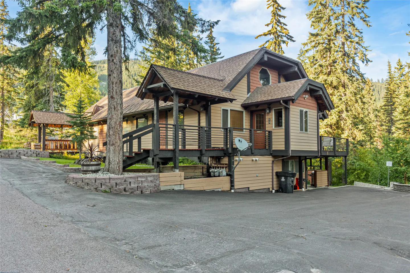 A tan two-story house with dark brown trim and a deck, surrounded by tall green trees.