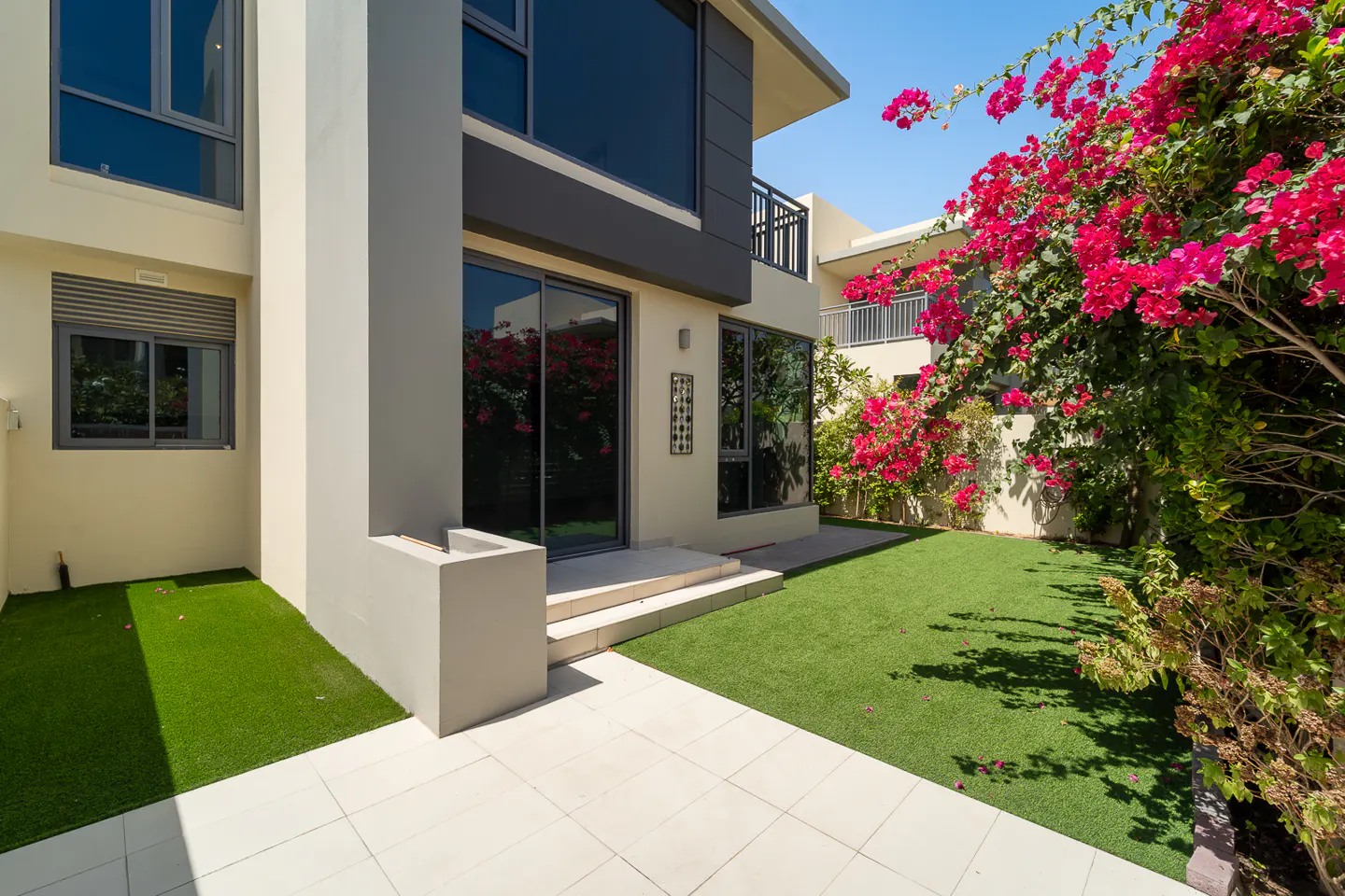 Exterior view of a modern two-story house with a patio, green lawn, and vibrant pink bougainvillea flowers.