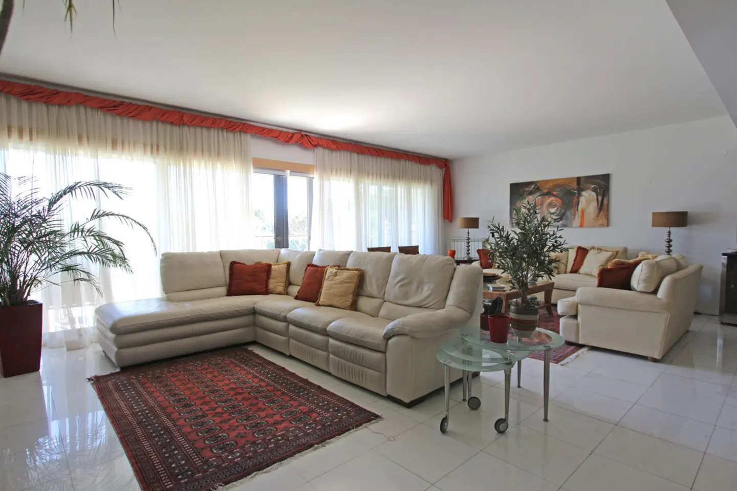 Bright living room with white tile floor, cream sofas with orange pillows, and a red patterned rug. Large windows with sheer curtains.