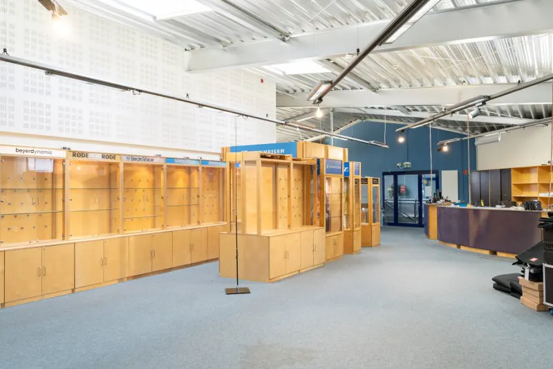 Interior view of a retail space with light wood display cases, blue carpet, and a high, white ceiling with industrial lighting.