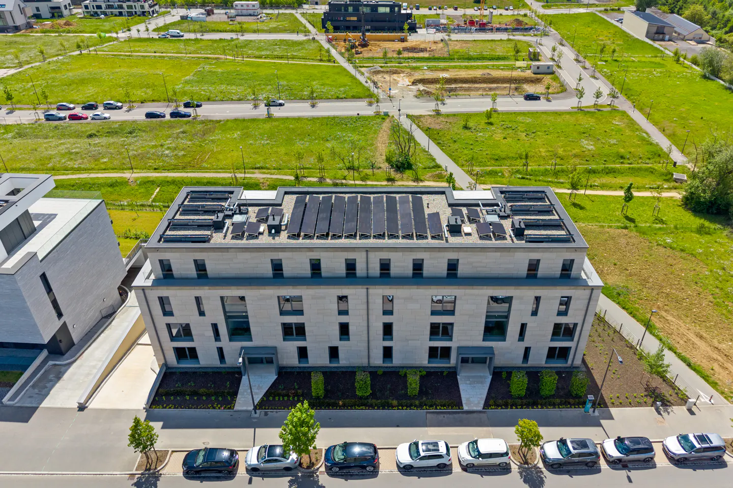 Aerial view of a modern, light-stone apartment building with solar panels on the roof, parked cars, and green space.