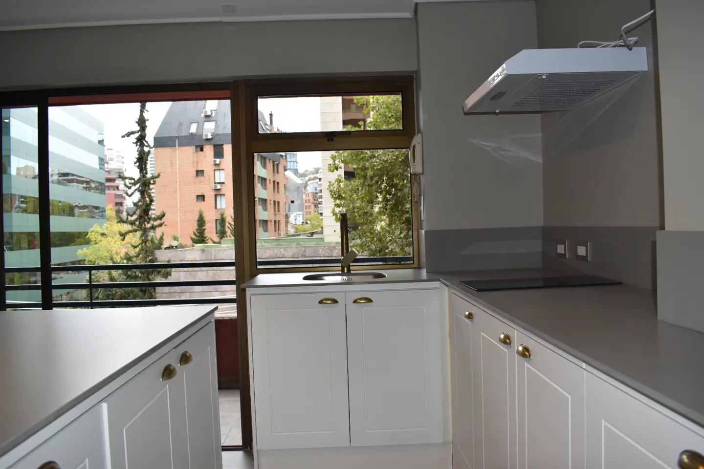 A modern kitchen with white cabinets, gold handles, and gray countertops. A window overlooks city buildings and trees.
