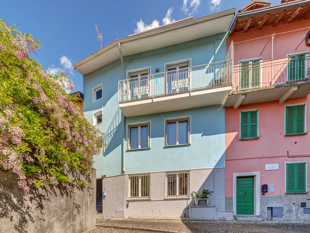 Exterior view of a light blue and pink building with a balcony and green shutters. Wisteria flowers cascade down a stone wall.