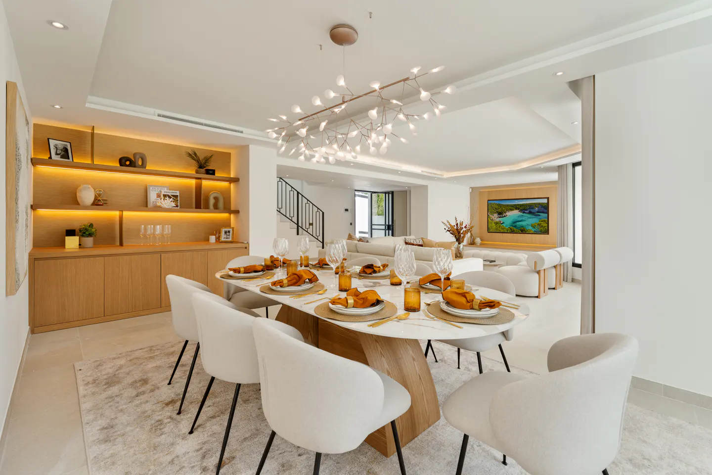 Bright, modern dining room with an oval table set for a meal. White chairs surround the table, with a unique chandelier overhead. A built-in shelf and TV are in the background.