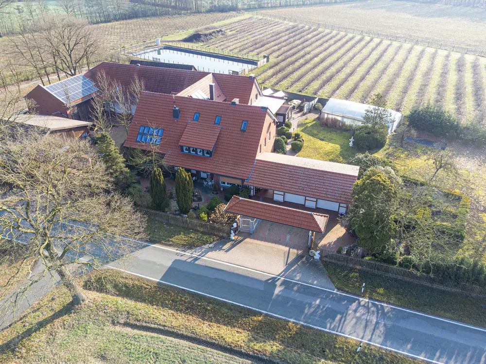 Aerial view of a red-roofed farmhouse with solar panels, a vineyard in the background, and a road in front.