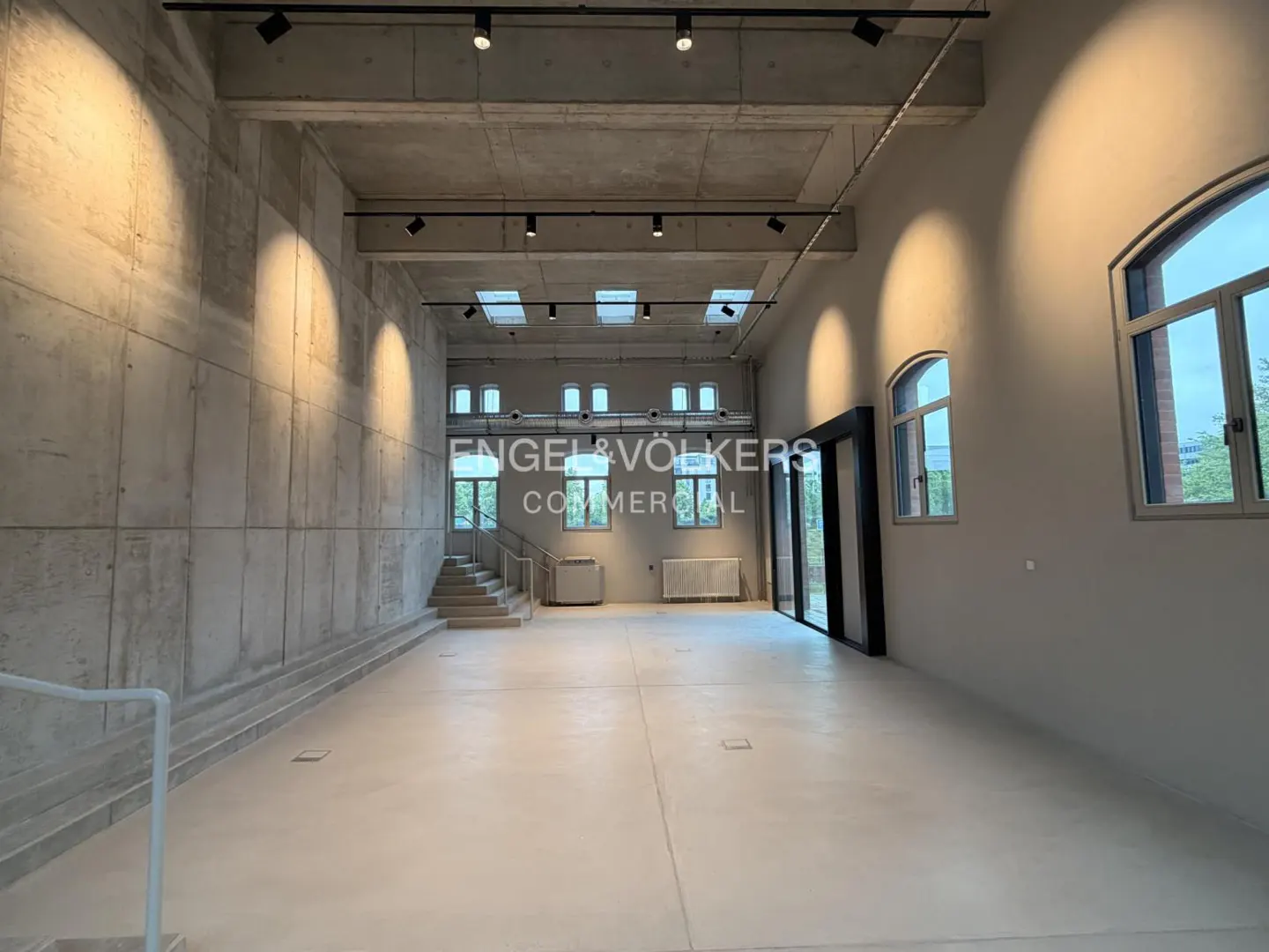 An empty commercial space with concrete walls, a light floor, and large windows. Track lighting is on the ceiling. Stairs lead to a raised area.