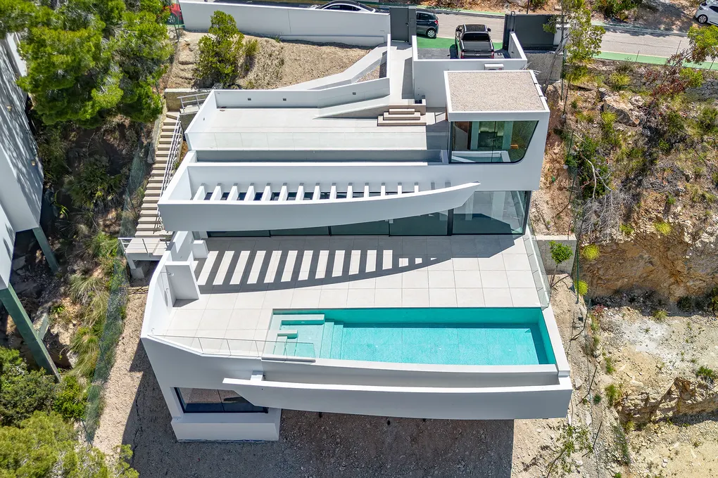 Aerial view of a modern white house with a turquoise pool, surrounded by trees and a hillside.