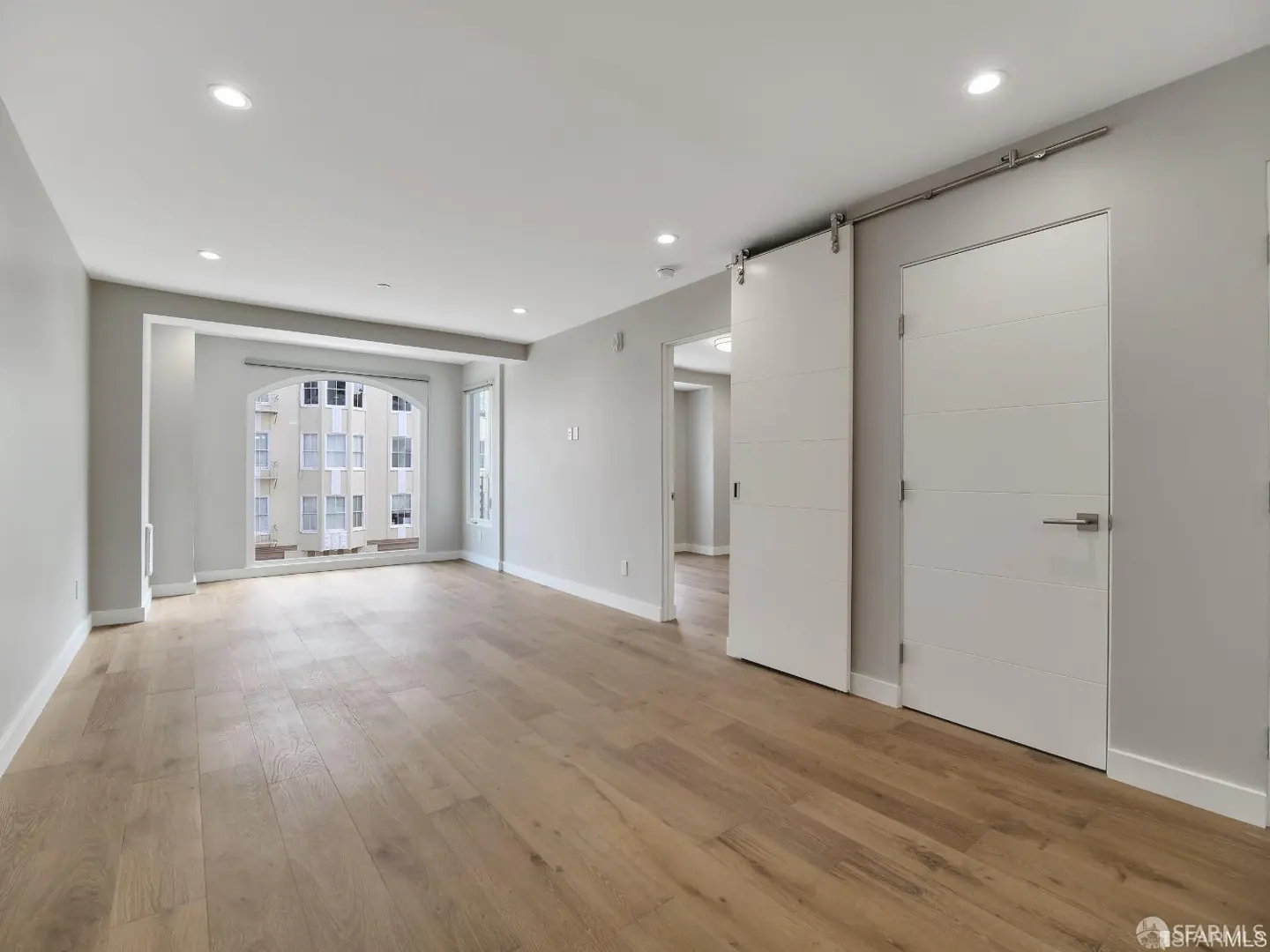 Bright, empty room with light wood floors, gray walls, and white trim. A large window shows a building exterior. A sliding barn door and a regular door are on the right.