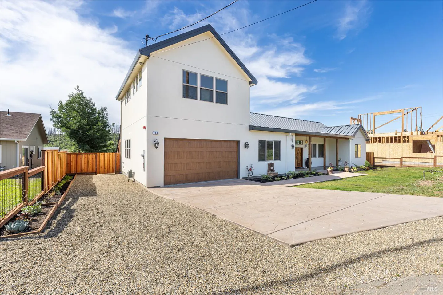 Two-story white house with a brown garage door, concrete driveway, and gravel yard. A wooden fence borders the property.