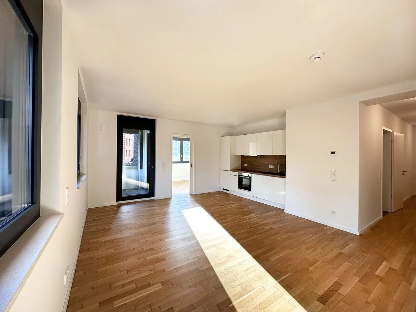 Modern open-plan kitchen with white cabinets, wood flooring, and a glass door leading to a balcony in a bright apartment.
