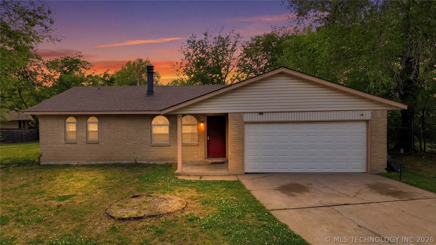 A single-story brick house with a brown roof, white garage door, and red front door at sunset.