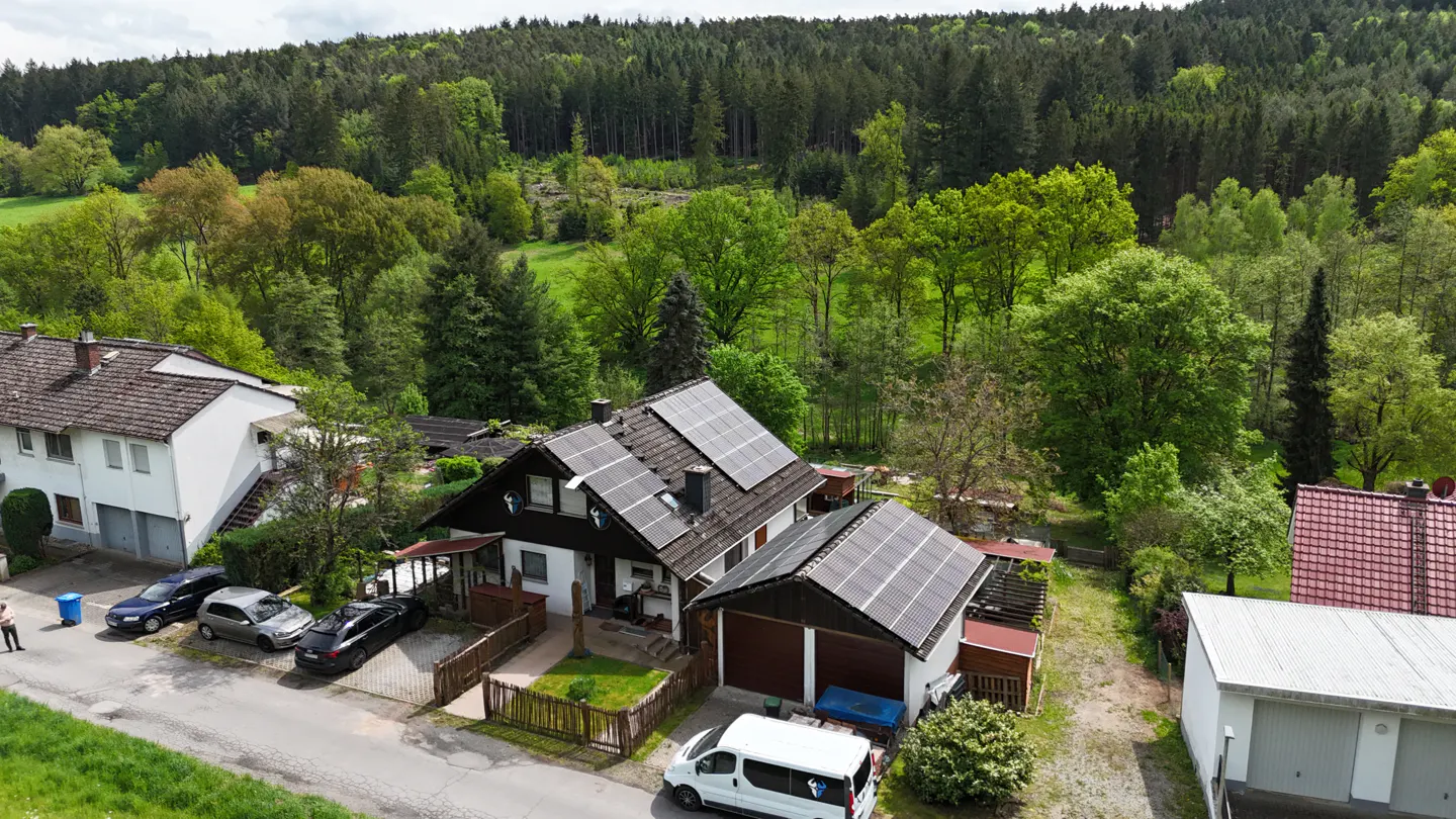 Aerial view of a two-story house with solar panels on the roof, surrounded by lush green trees and a forest in the background.