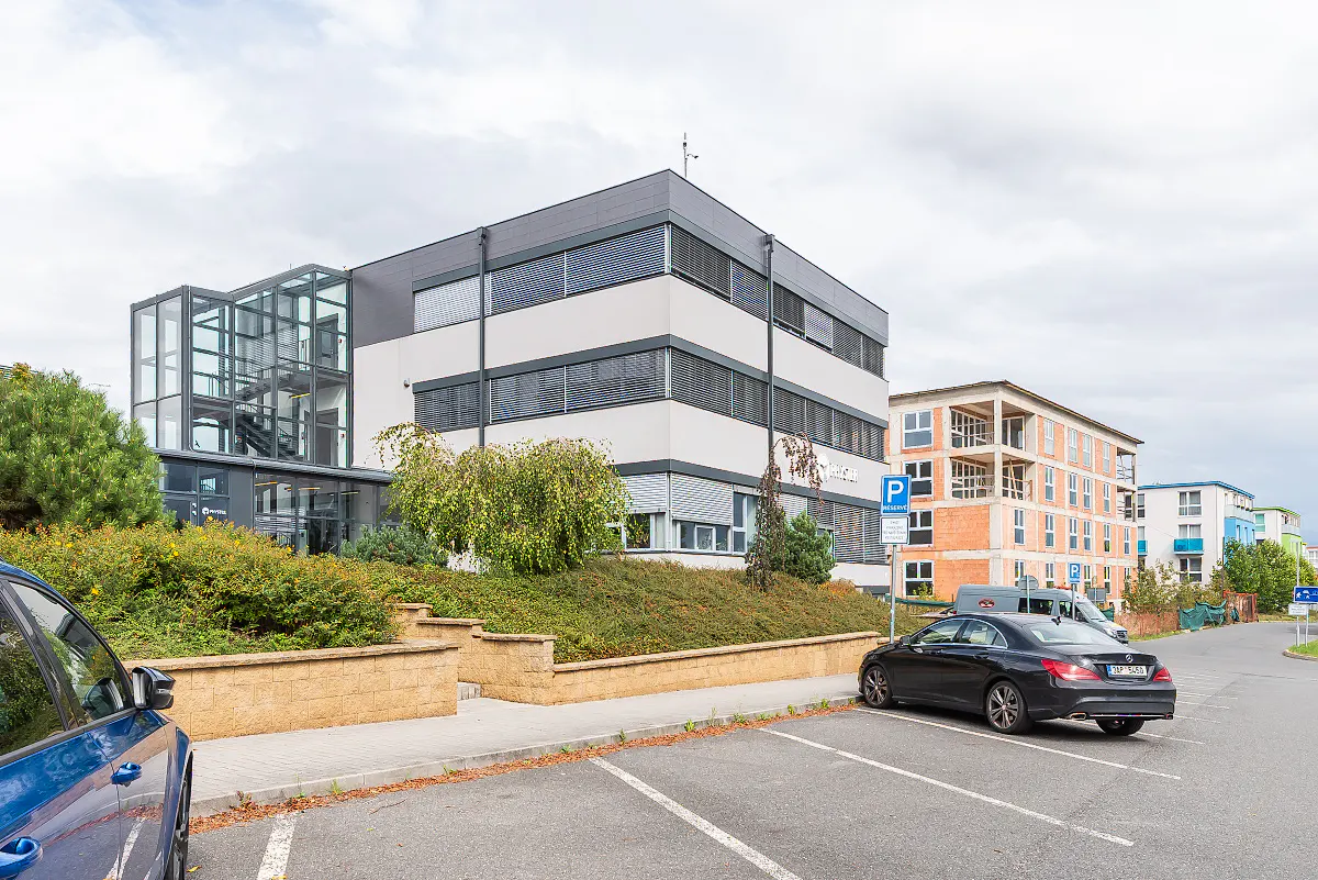 A modern, three-story office building with a glass staircase entrance and parking lot. A black car is parked in front.