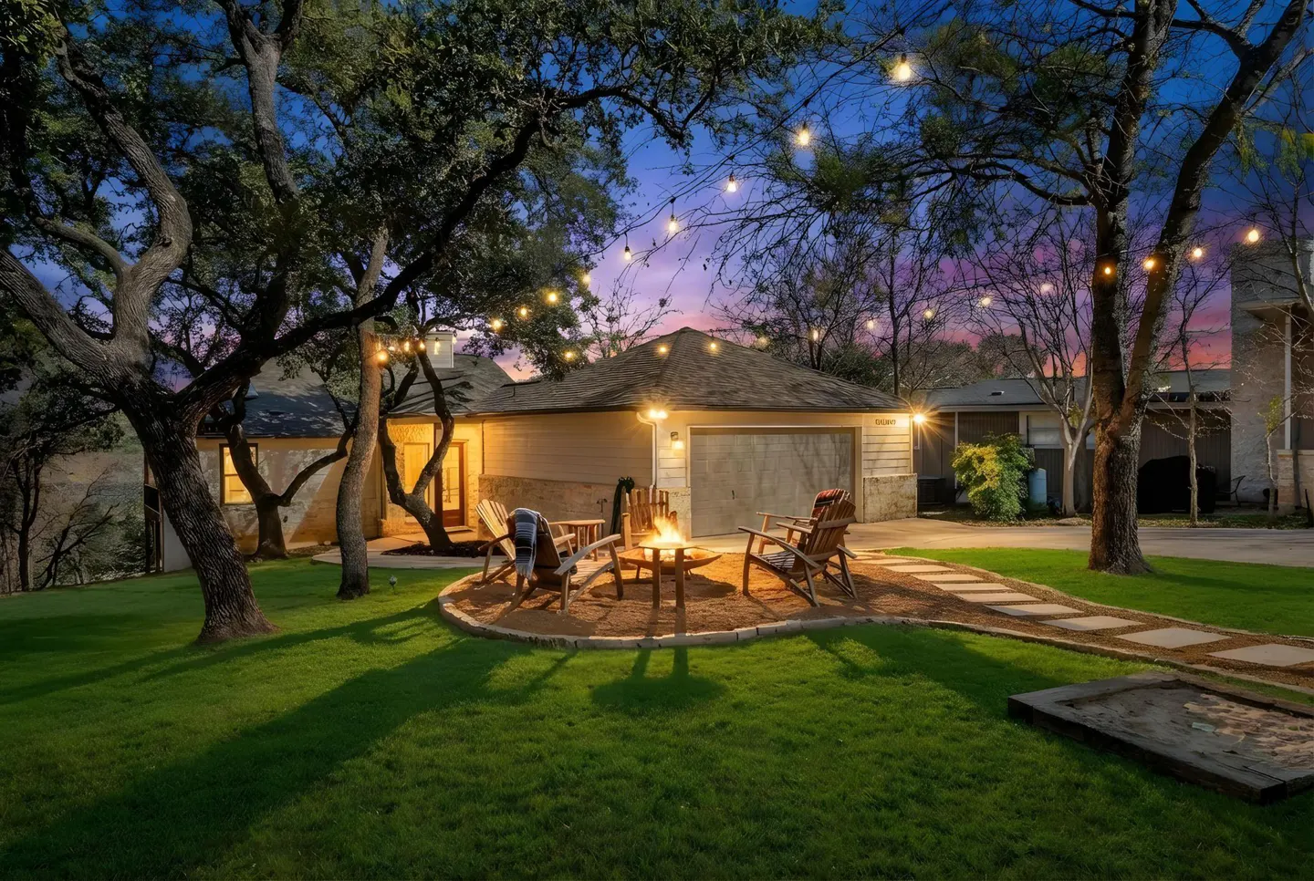 Outdoor patio with a fire pit and chairs, string lights, green grass, and a house in the background at dusk.