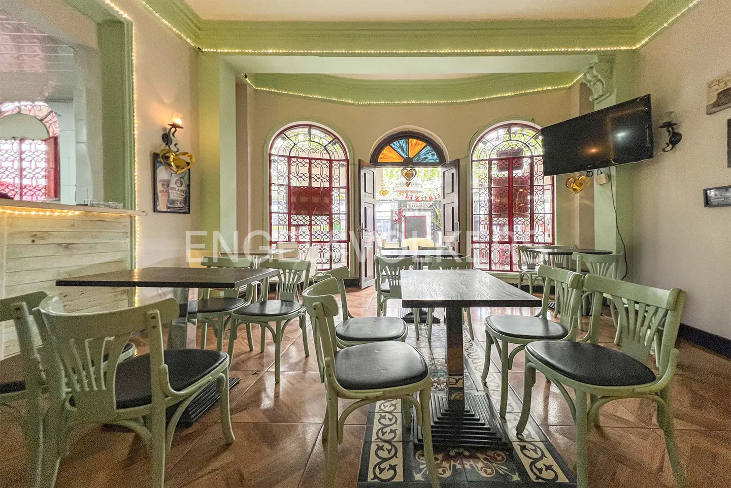 Interior view of a restaurant with green chairs and dark tables, arched windows with red frames, and a TV on the wall.