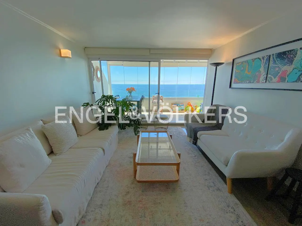 Living room with white sofas, glass table, and ocean view through a glass balcony.