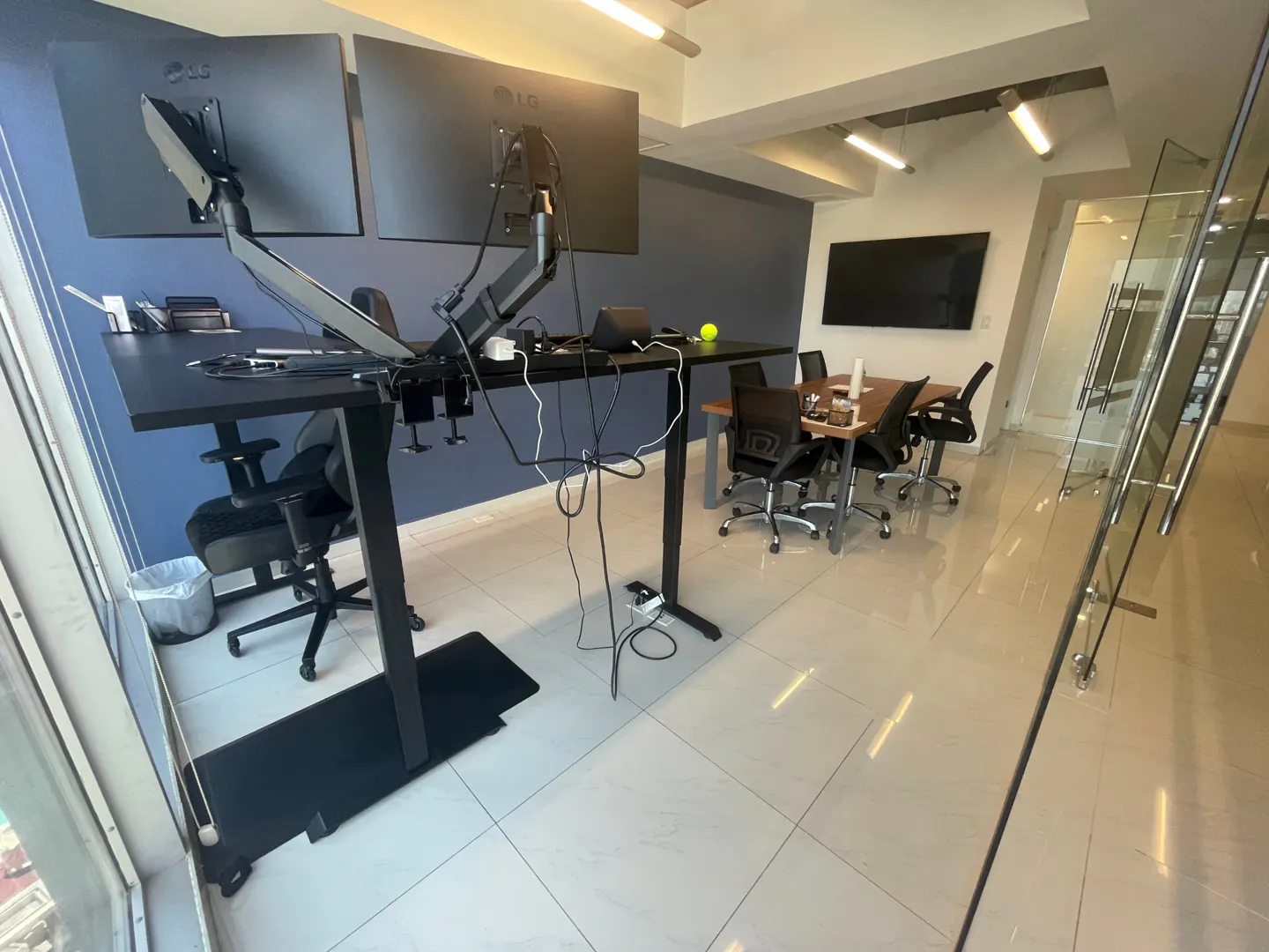 Office space with two black adjustable desks, dual monitors, and a conference table with chairs. The floor is tiled and there is a glass wall.
