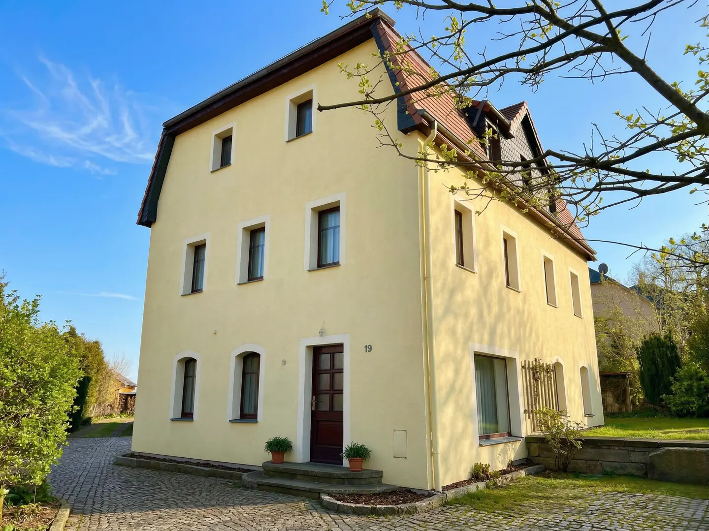 Two-story yellow house with a red tile roof and brown trim, set against a blue sky. The house has a brown front door and is surrounded by greenery.