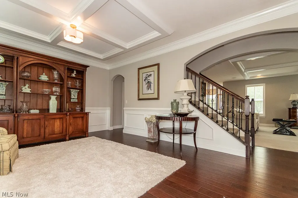 A foyer with dark wood floors, a cream rug, and a dark wood hutch filled with vases. A staircase is visible through an archway.
