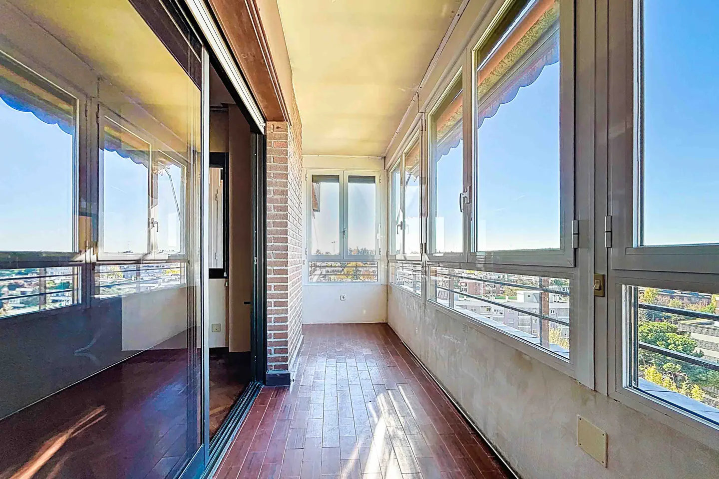 Enclosed balcony with wood floors, brick accent wall, and large windows overlooking a city on a sunny day.