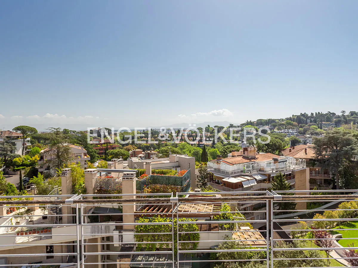 View from a balcony with a metal railing overlooking rooftops, trees, and a blue sky. Engel & Völkers logo is visible in the background.