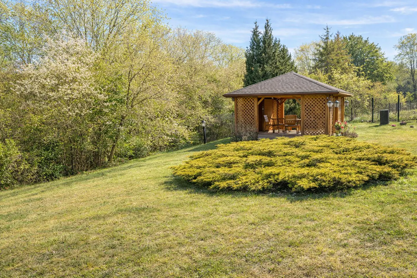 A wooden gazebo sits on a green lawn, surrounded by trees and bushes.