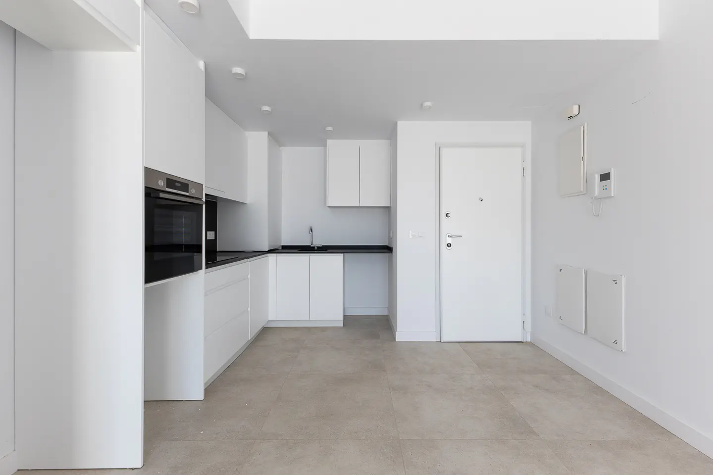 Minimalist kitchen with white cabinets, black countertops, built-in oven, and light grey tiled floors next to a white door.