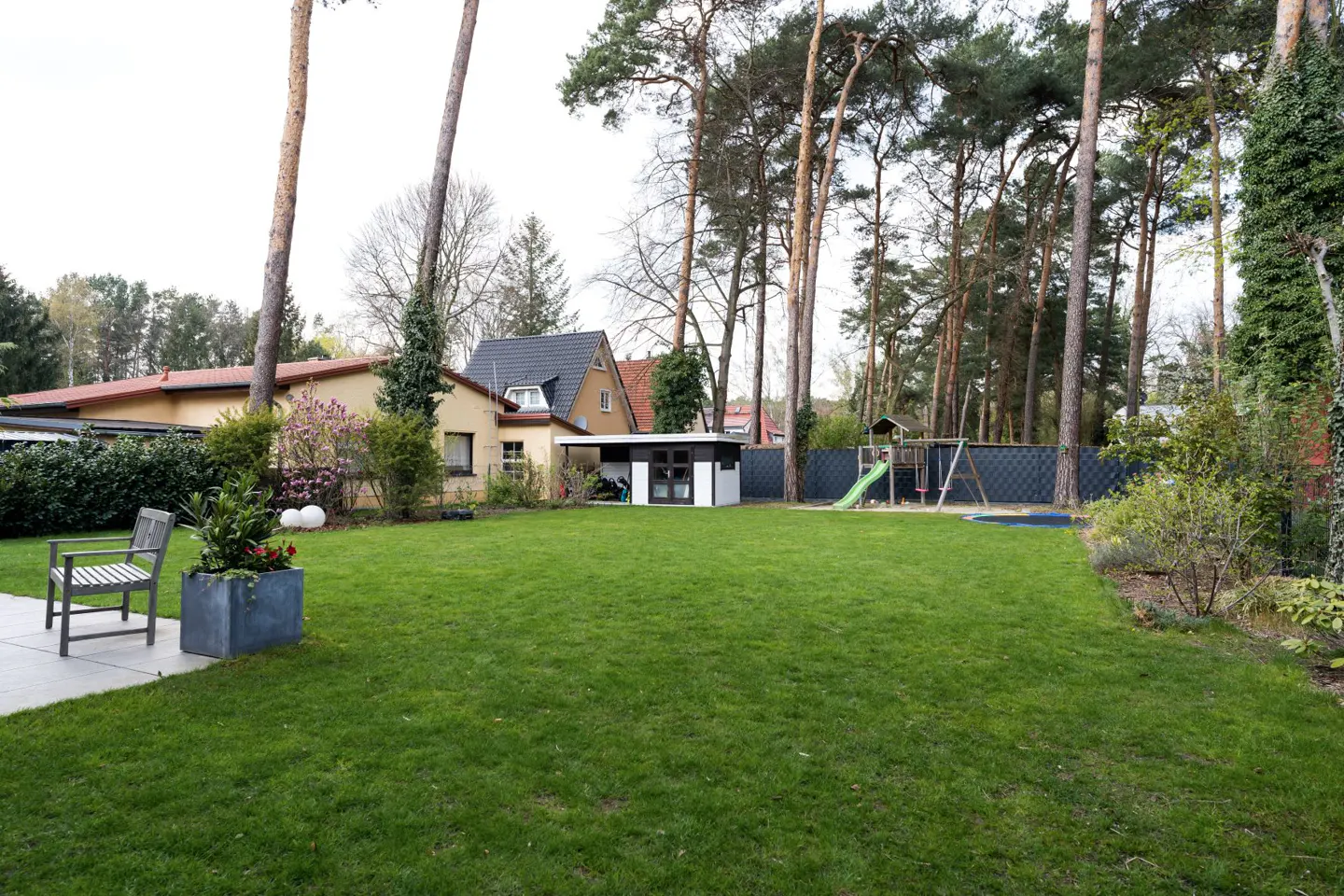A lush green lawn stretches to a house, shed, and playground set, framed by tall trees. A bench and planter sit on a patio in the foreground.