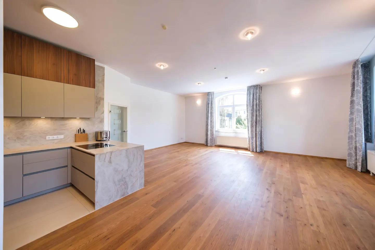 A bright, open-concept apartment with hardwood floors, a modern kitchen with gray cabinets, and large windows with patterned curtains.