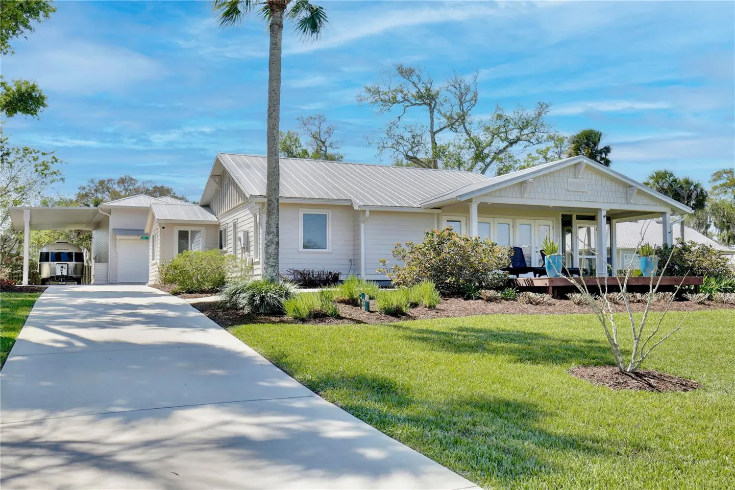 Front exterior of a one-story beige house with a metal roof, a carport, and a green lawn on a sunny day.
