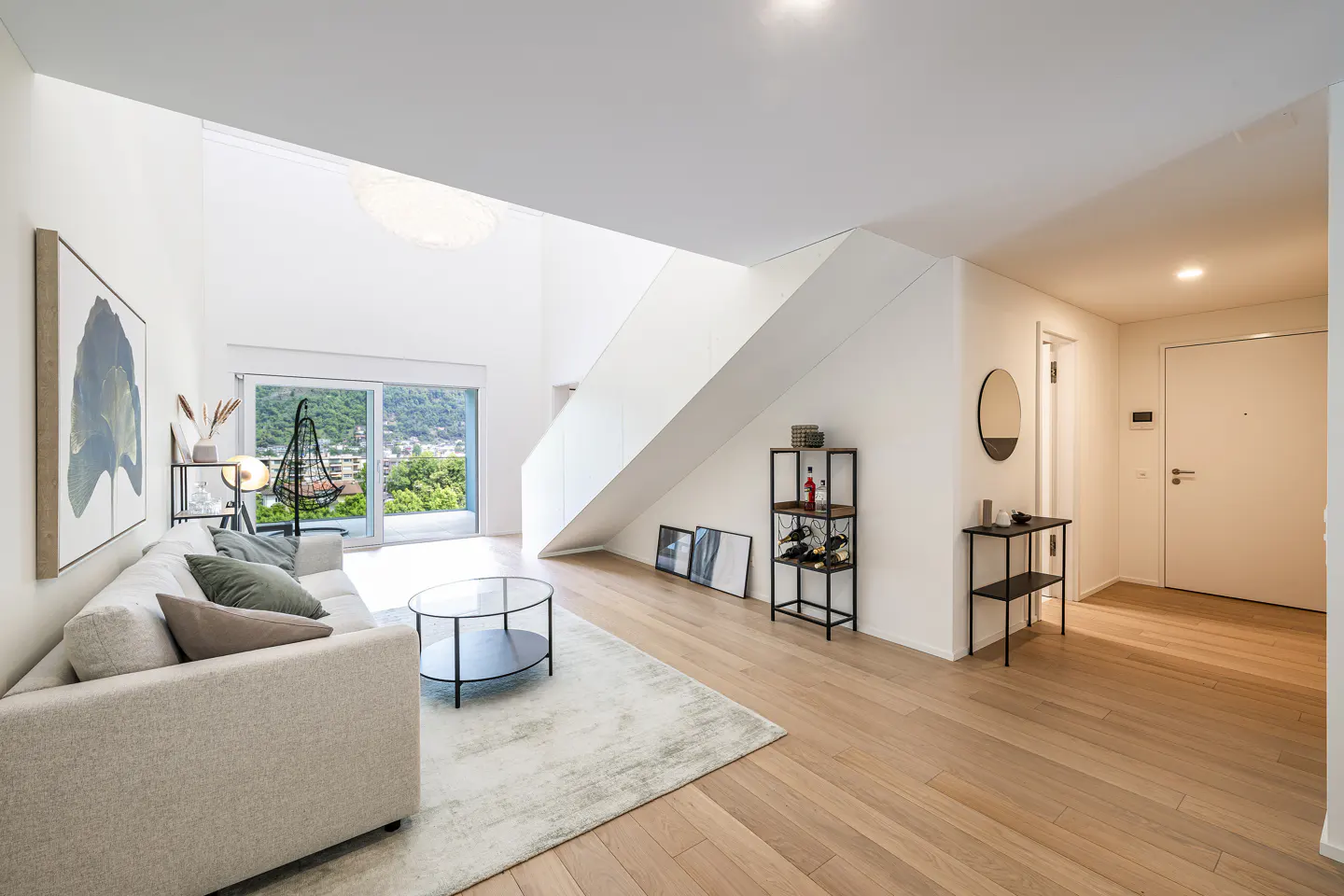 Bright, modern living room with white walls, light wood floors, and a beige sofa. A staircase is visible in the background, along with a view of greenery through a sliding glass door.