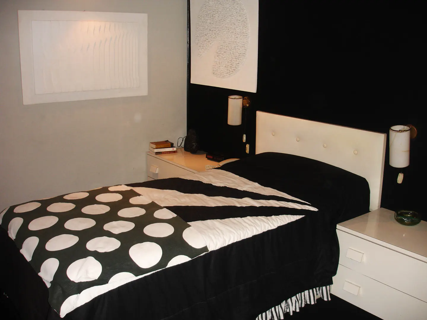 Bedroom with a bed covered in a black and white polka dot and striped comforter. White nightstands flank the bed with white headboard. Black wall behind the bed.