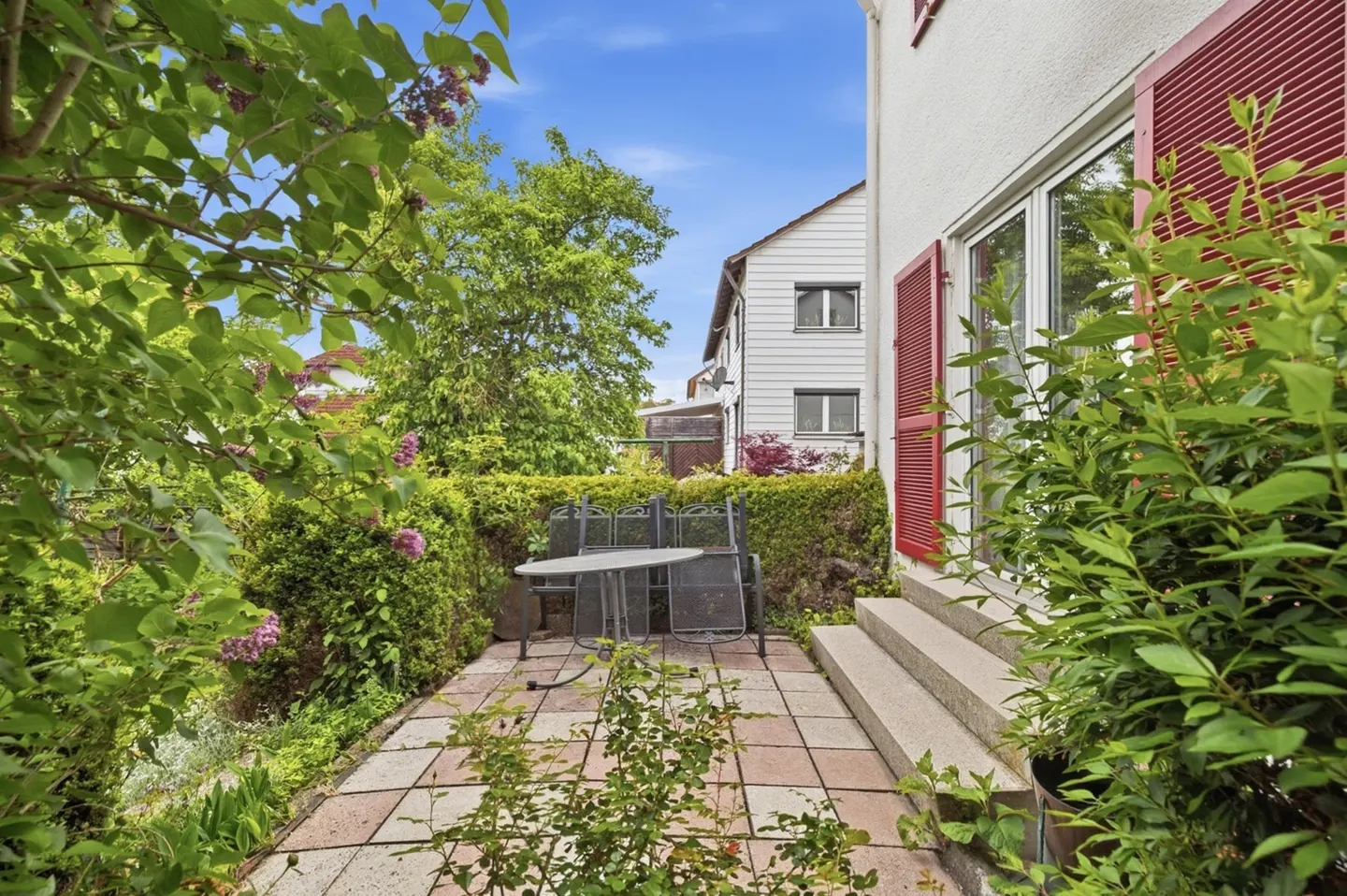 A patio with a table and chairs is surrounded by greenery, next to a white house with red shutters.