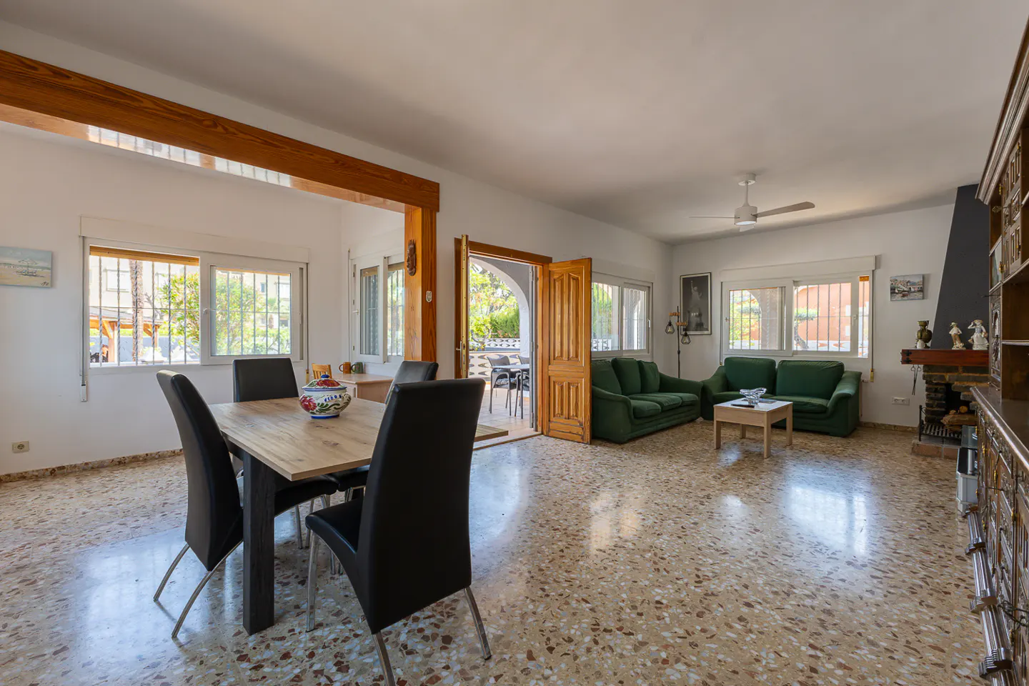 Bright living room with dining table and green sofas. The floor is speckled. A fireplace is on the right. Windows and doors let in natural light.