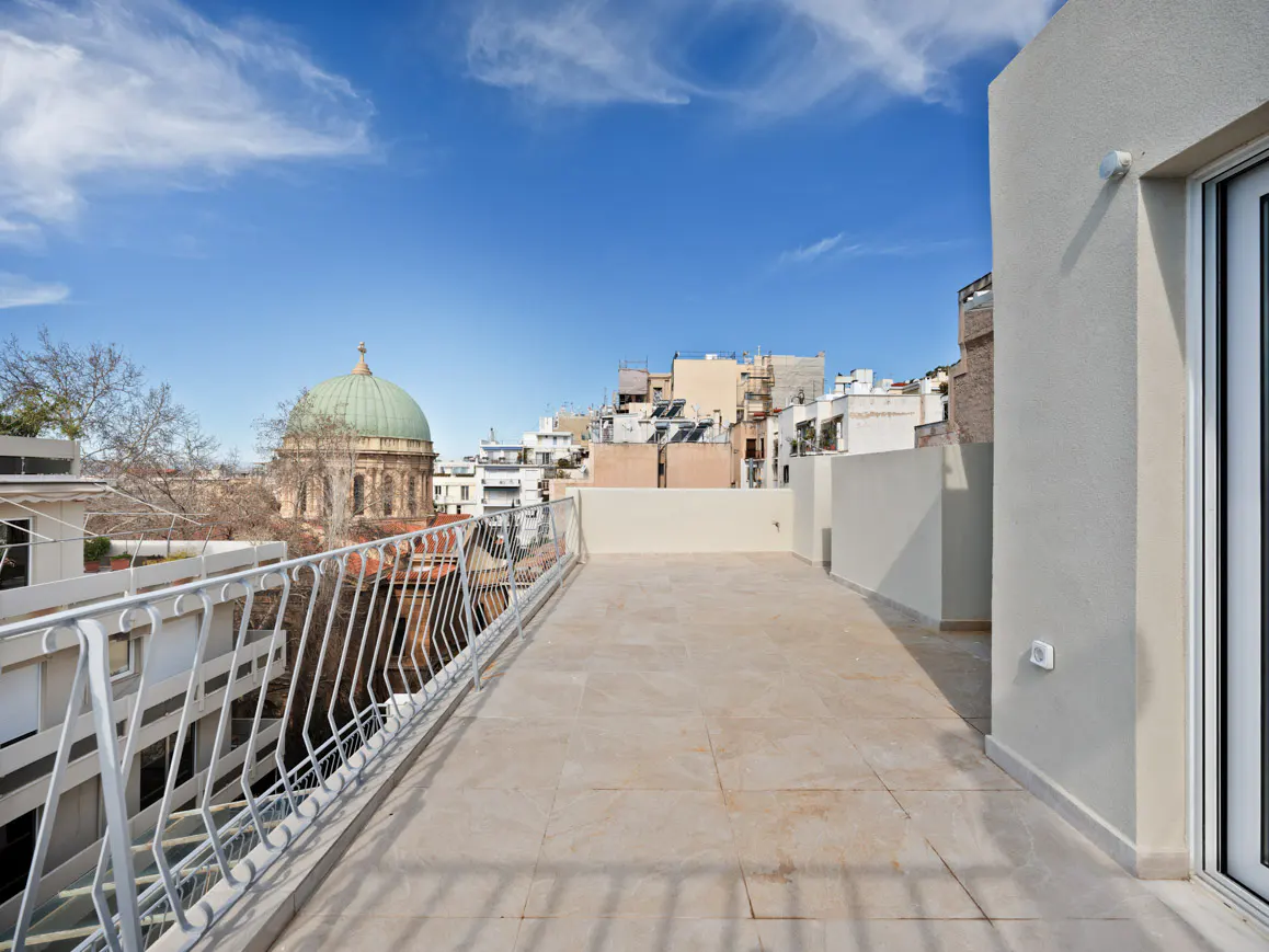 Wide, tiled rooftop terrace with white railings, overlooking a city with a domed building under a blue sky.