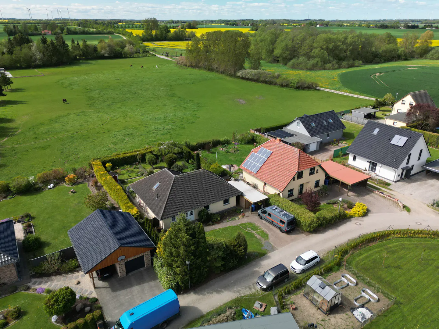 Aerial view of houses with solar panels, cars, and green fields under a blue sky.