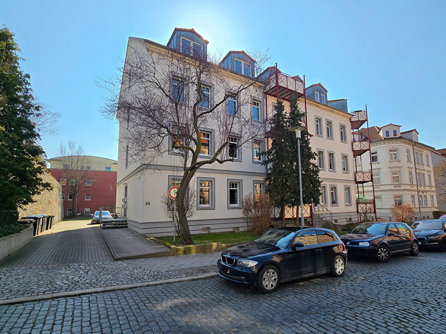 A three-story white apartment building with a red fire escape and parked cars on a cobblestone street.