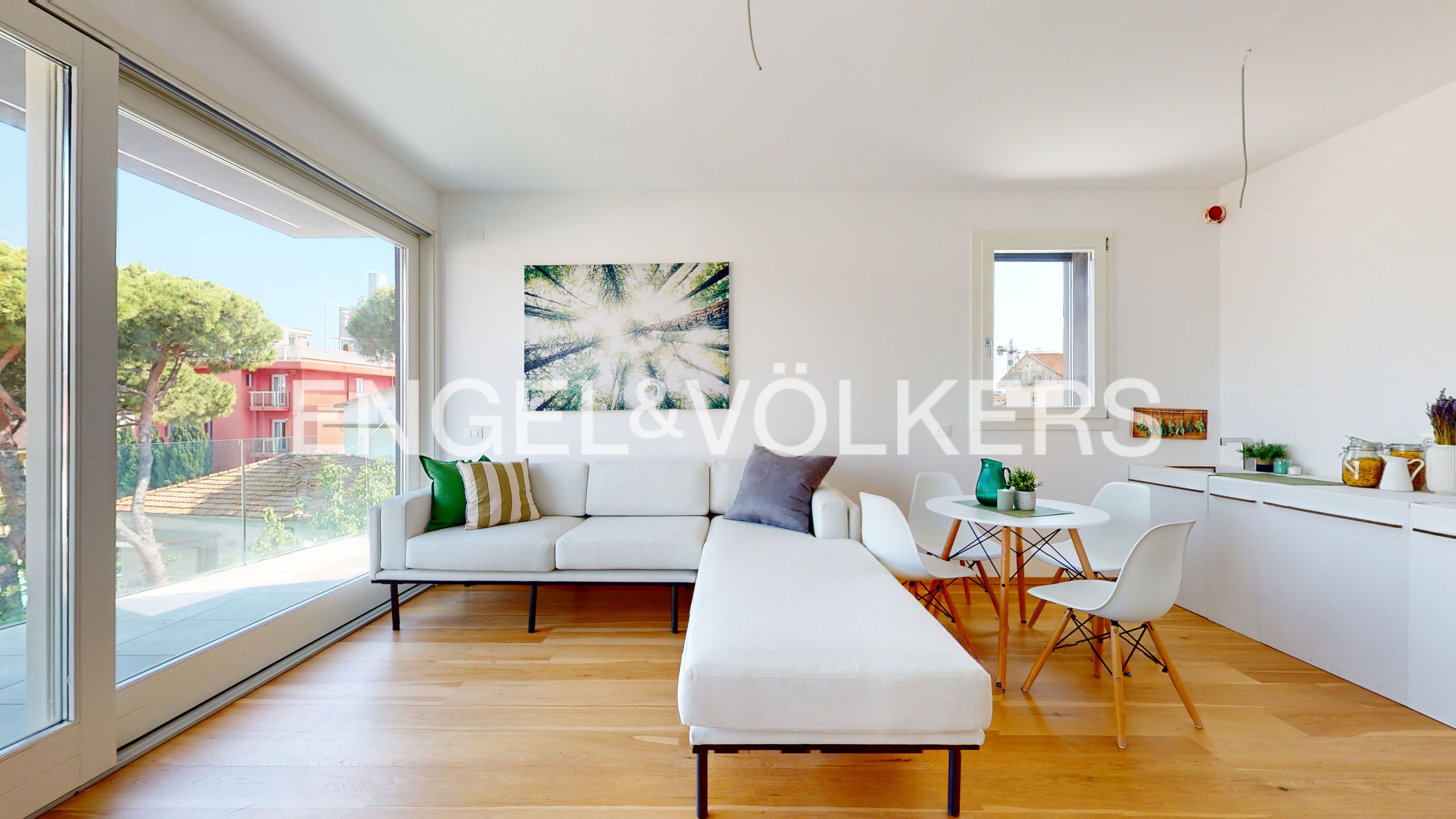 Bright, modern living room with white walls, wood floors, and a large window. A white sectional sofa faces a round table with white chairs. An abstract tree art piece hangs on the wall.