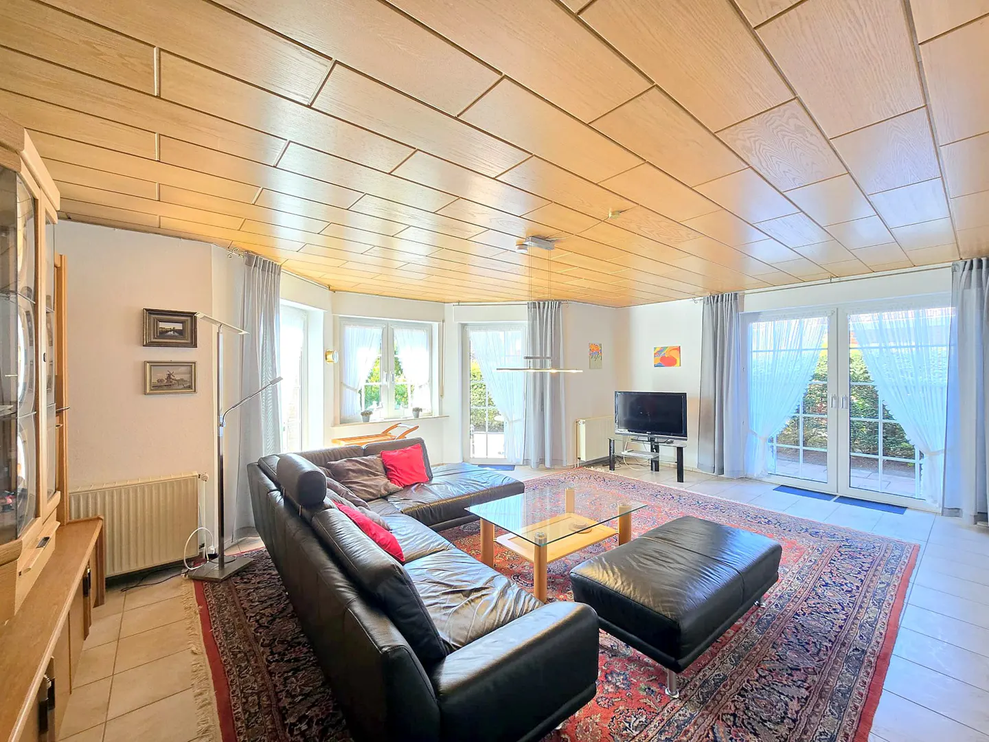 Living room with wood-paneled ceiling, black leather sectional sofa, glass coffee table, and red patterned rug. French doors lead to a garden.