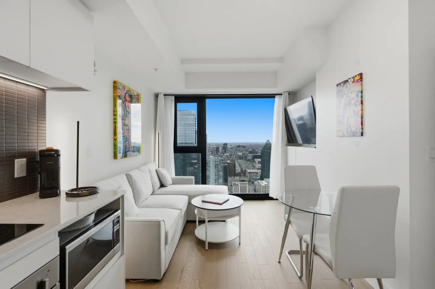 Bright, modern condo interior with white sectional sofa, glass table, and city view from a large window.