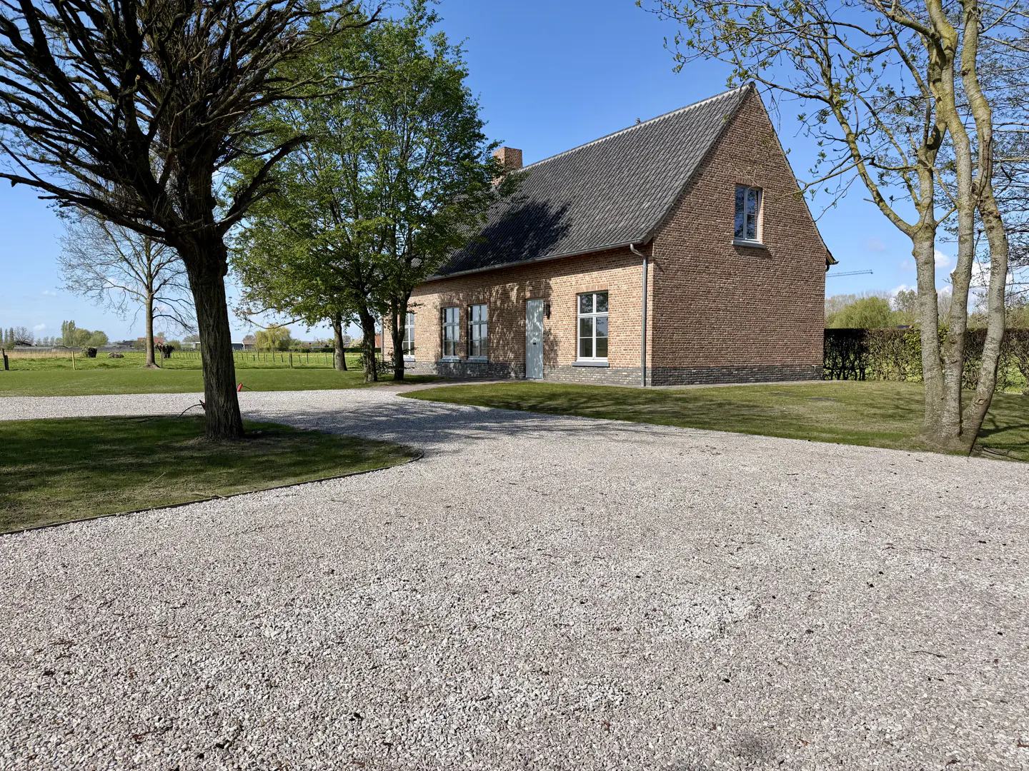 Brick house with a gray roof and white trim, surrounded by green grass and trees, with a gravel driveway.