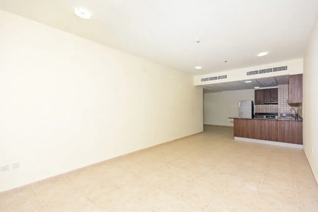 An empty room with beige tile flooring and off-white walls, leading to a kitchen with brown cabinets and stainless steel appliances.