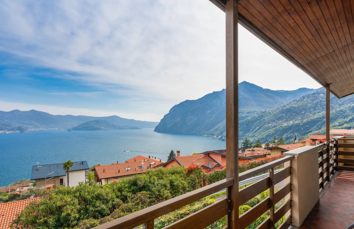 View from a balcony with wooden railings overlooking a lake, mountains, and houses with red tile roofs.