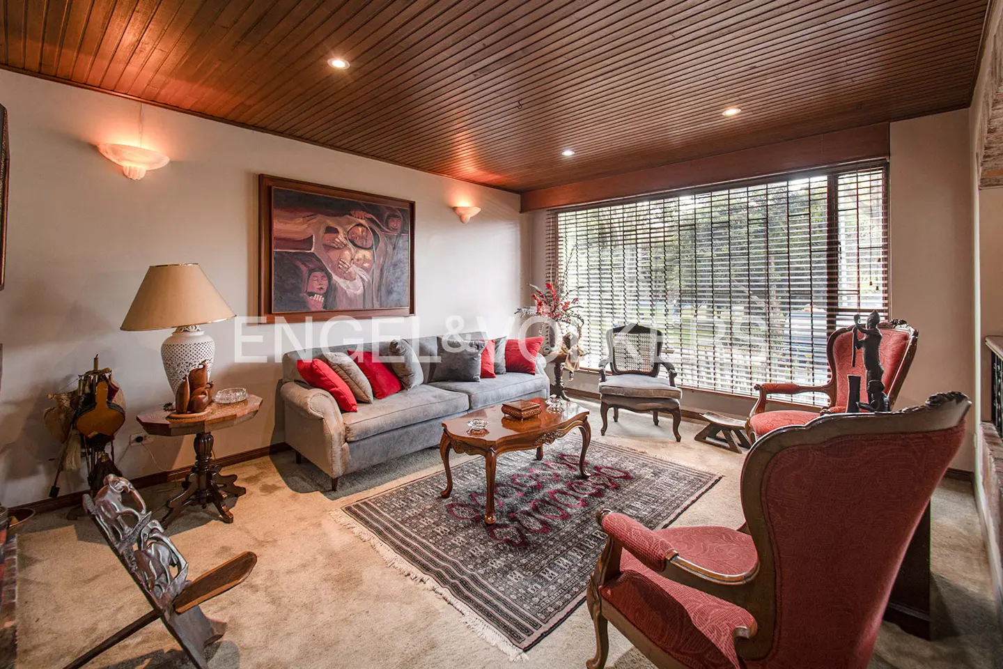 Living room with wood ceiling, large window with blinds, gray sofa with red pillows, and red patterned chairs.