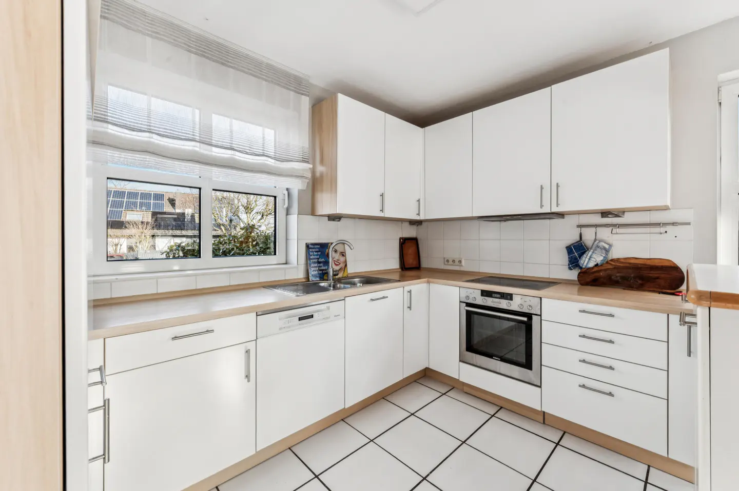 Bright kitchen with white cabinets, light wood countertops, and white tile floor. A window overlooks a neighborhood with solar panels.
