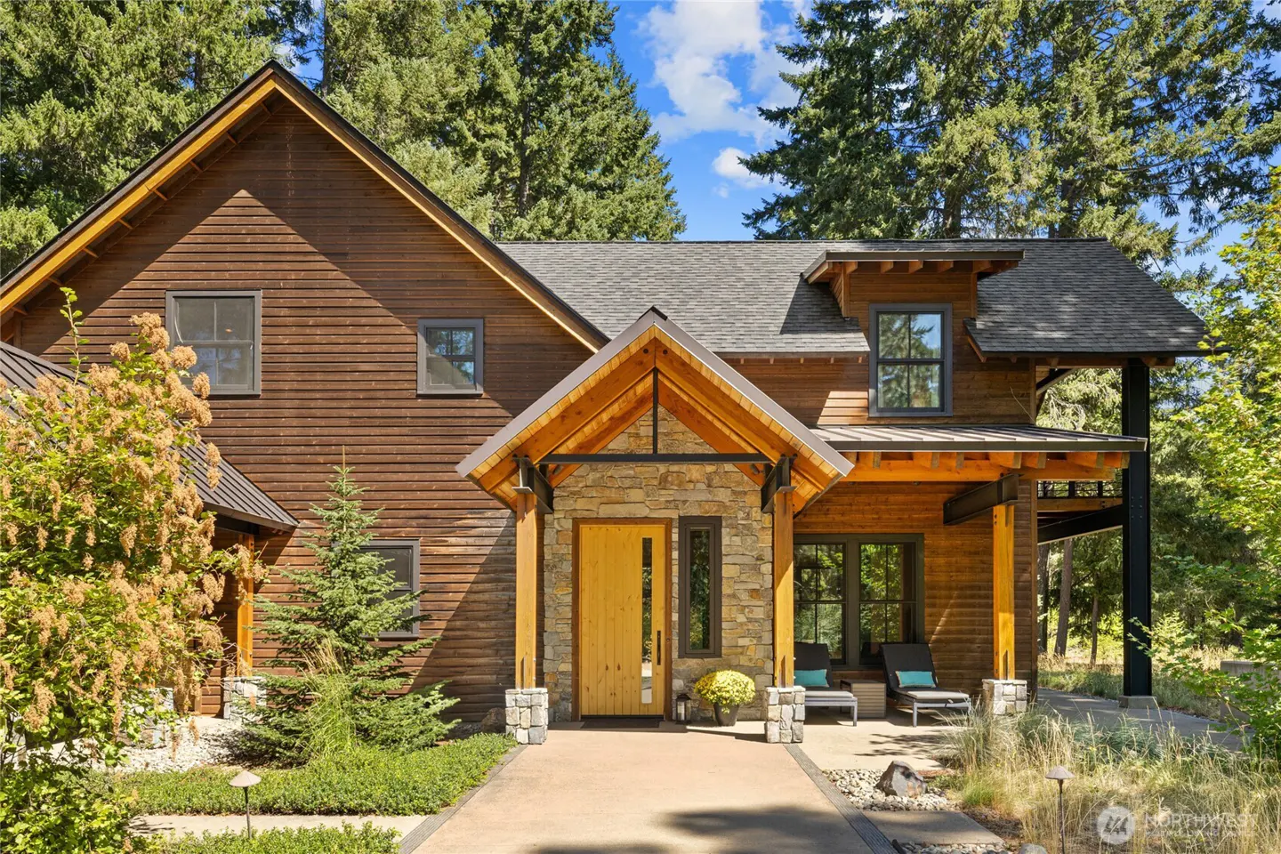 A two-story wood-sided home with a stone entryway, light wood door, and a covered porch with lounge chairs in a forest.