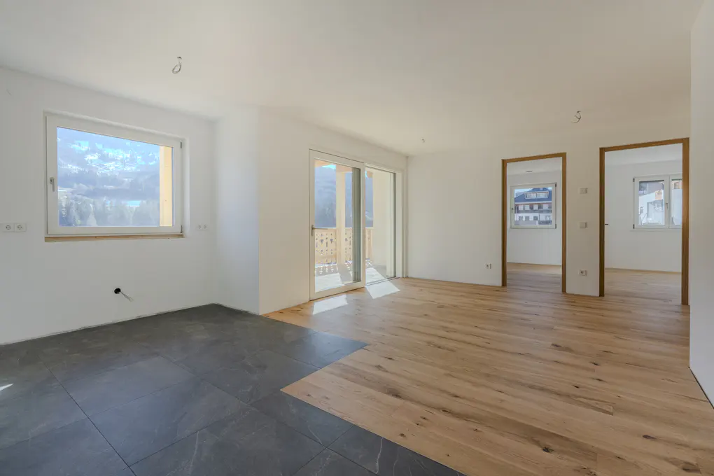 Bright, empty room with white walls, wood floors, and a window with a mountain view. A sliding glass door leads to a balcony.