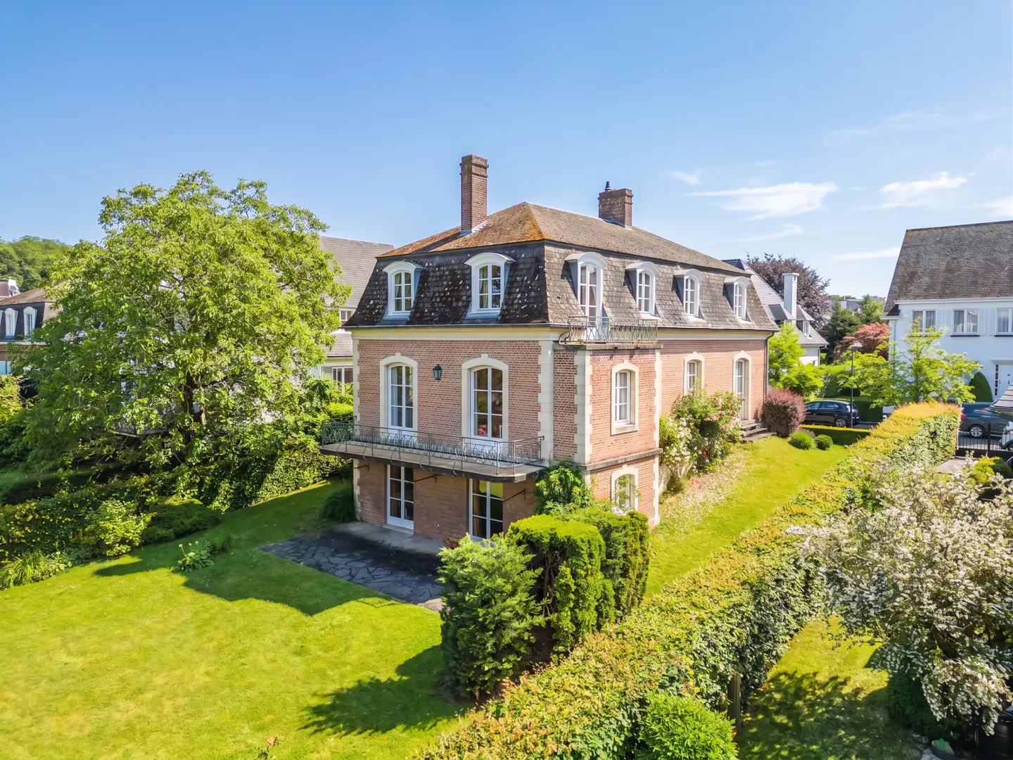 A two-story brick mansion with a mansard roof, balcony, and dormer windows, set in a lush green garden on a sunny day.