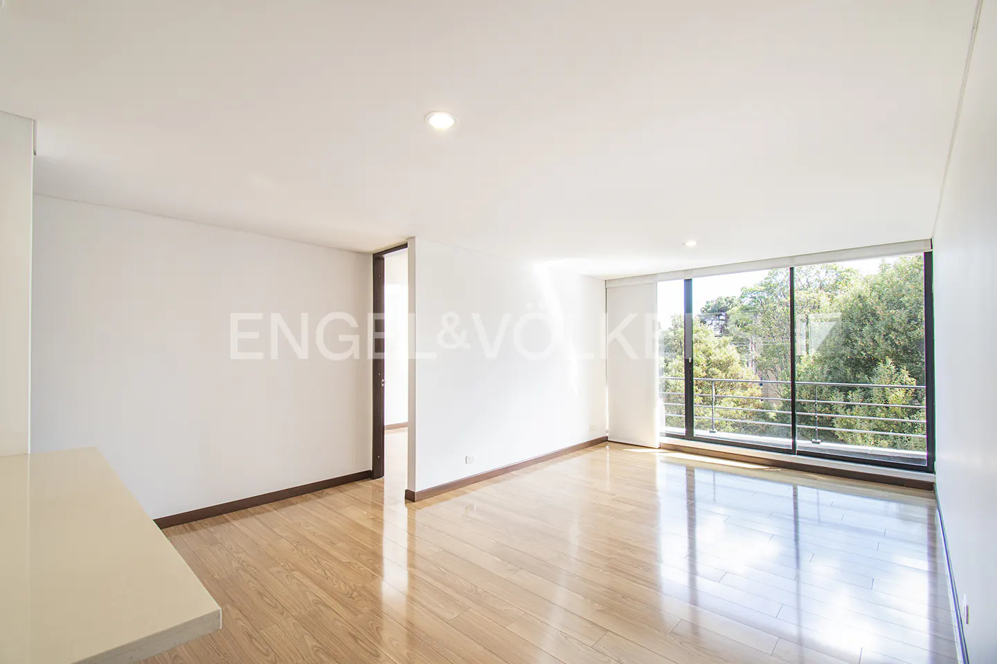 Bright, empty room with wood floors, white walls, and a large window showing trees. Engel & Völkers logo on the wall.